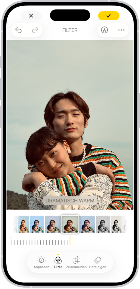 Der Bildschirm „Bearbeiten“ mit einem Foto in der Mitte. Unten auf dem Bildschirm sind von links nach rechts die Tasten „Anpassen“, „Filter“, „Zuschneiden“ und „Bereinigen“ zu sehen. Die Taste „Filter“ ist ausgewählt. Unter dem Foto befinden sich verschiedene Filteroptionen. „Dramatisch warm“ ist ausgewählt und ein Schieberegler ermöglicht die Anpassung der Filterintensität. Die Taste „Abbrechen“ befindet sich oben links und die Taste „Fertig“ oben rechts.