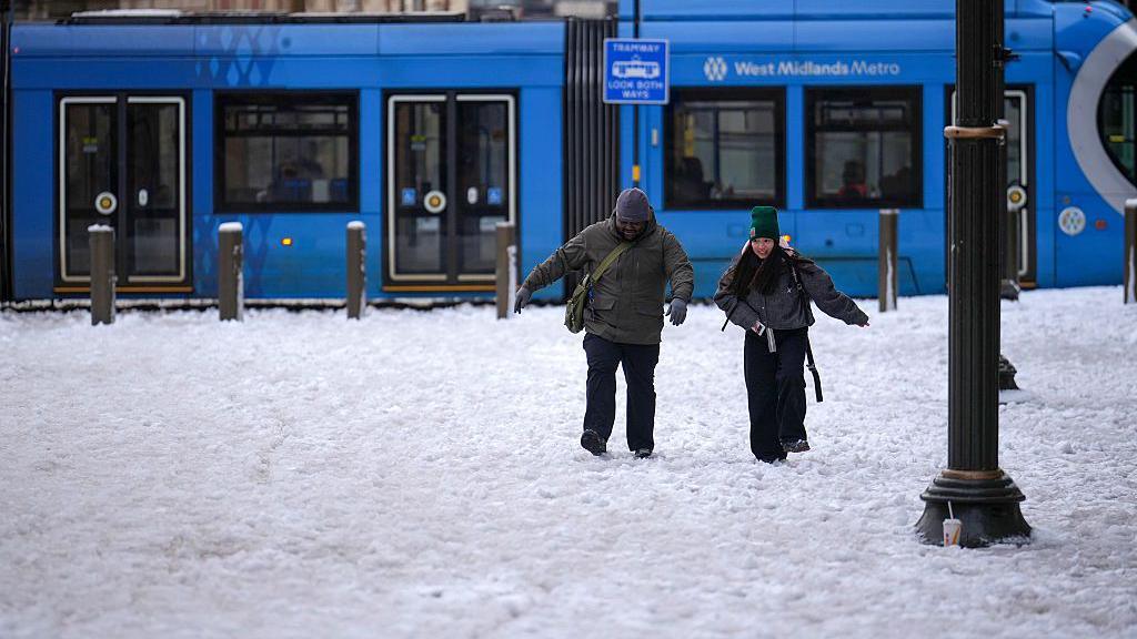 Two people walk through slushy snow away from a tram labelled 'West Midlands Metro'