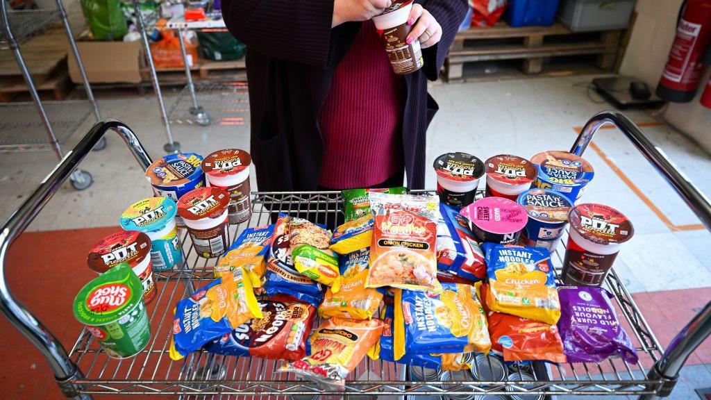 Picture of a trolly with multiple packs of instant noodles and pasta on it. We can see someone standing behind the trolley opening a packet but their face is not visible.