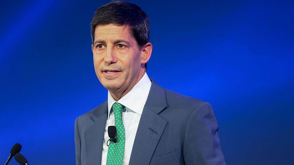 Kevin Warsh, wearing a blue suit and green tie stands in front of a podium with a blue background behind him