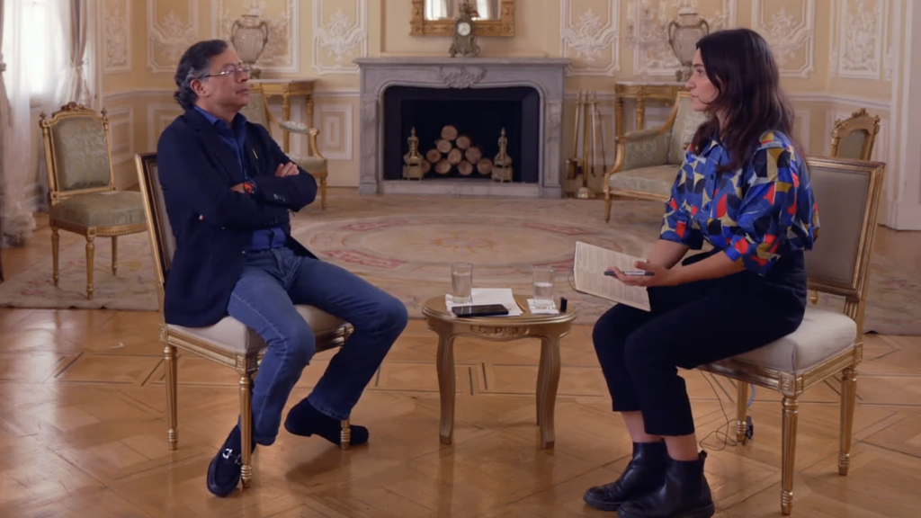 Gustavo Petro and Ione Wells sit facing each other in an ornate room with parquet flooring and cream-colored walls decorated with intricate moldings