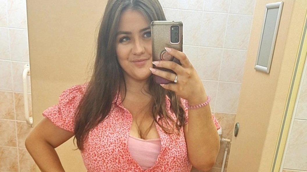 Melissa Ashcroft wearing a pink patterned dress taking a mirror selfie. She has straight brown hair and is smiling.