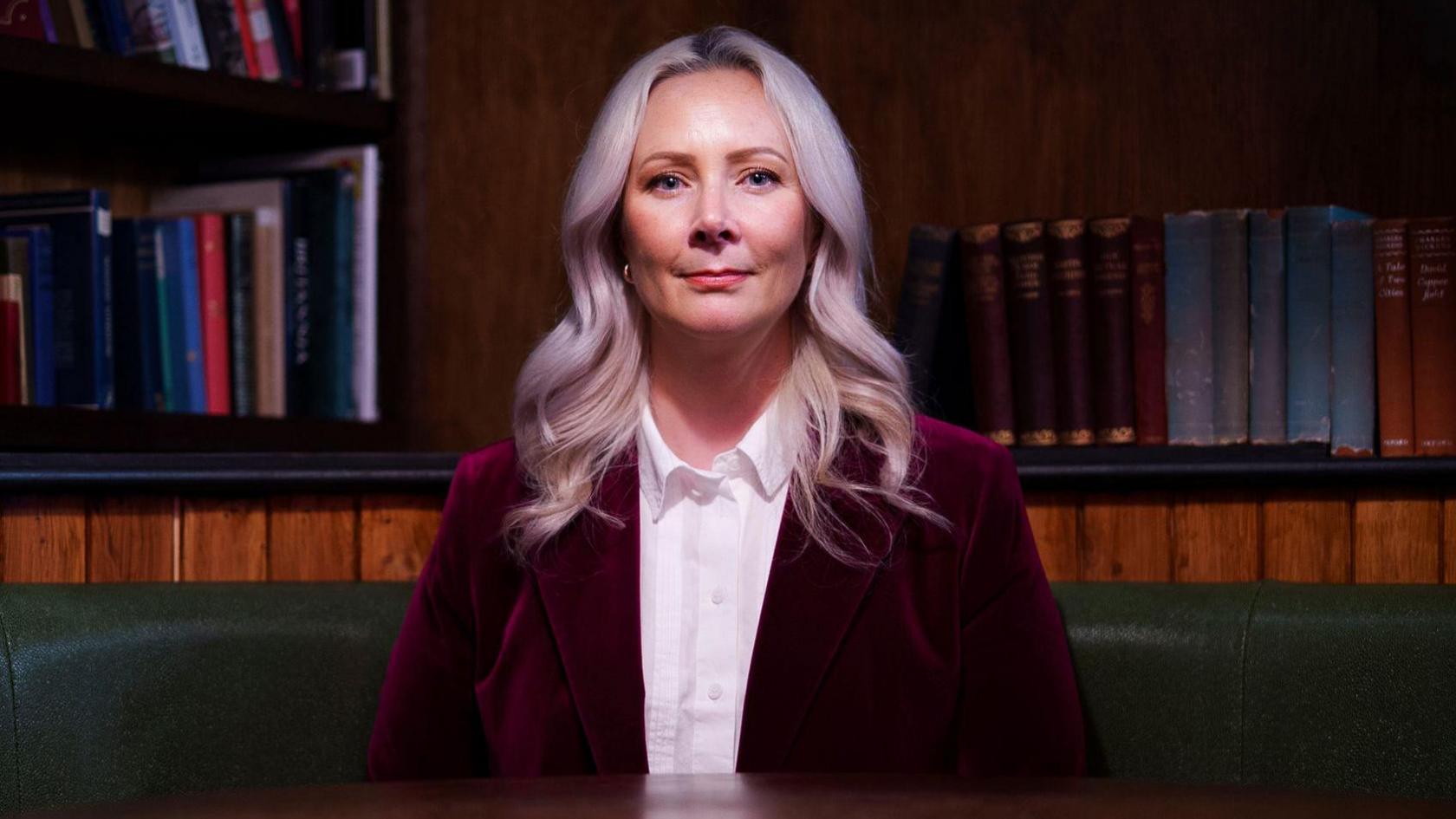 A woman with long, wavy, light hair sitting in a green‑backed booth, wearing a white shirt and dark red velvet blazer, with bookshelves filled with books in the background