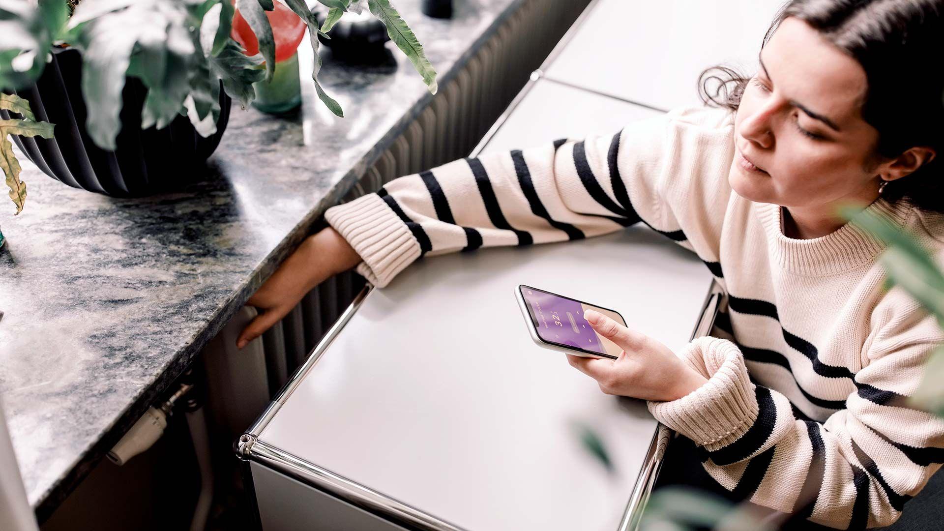 A high angle view of young woman wearing a white and black striped jumper who is checking the temperature of a radiator next to a window. She is looking at a heating app on her smart phone.