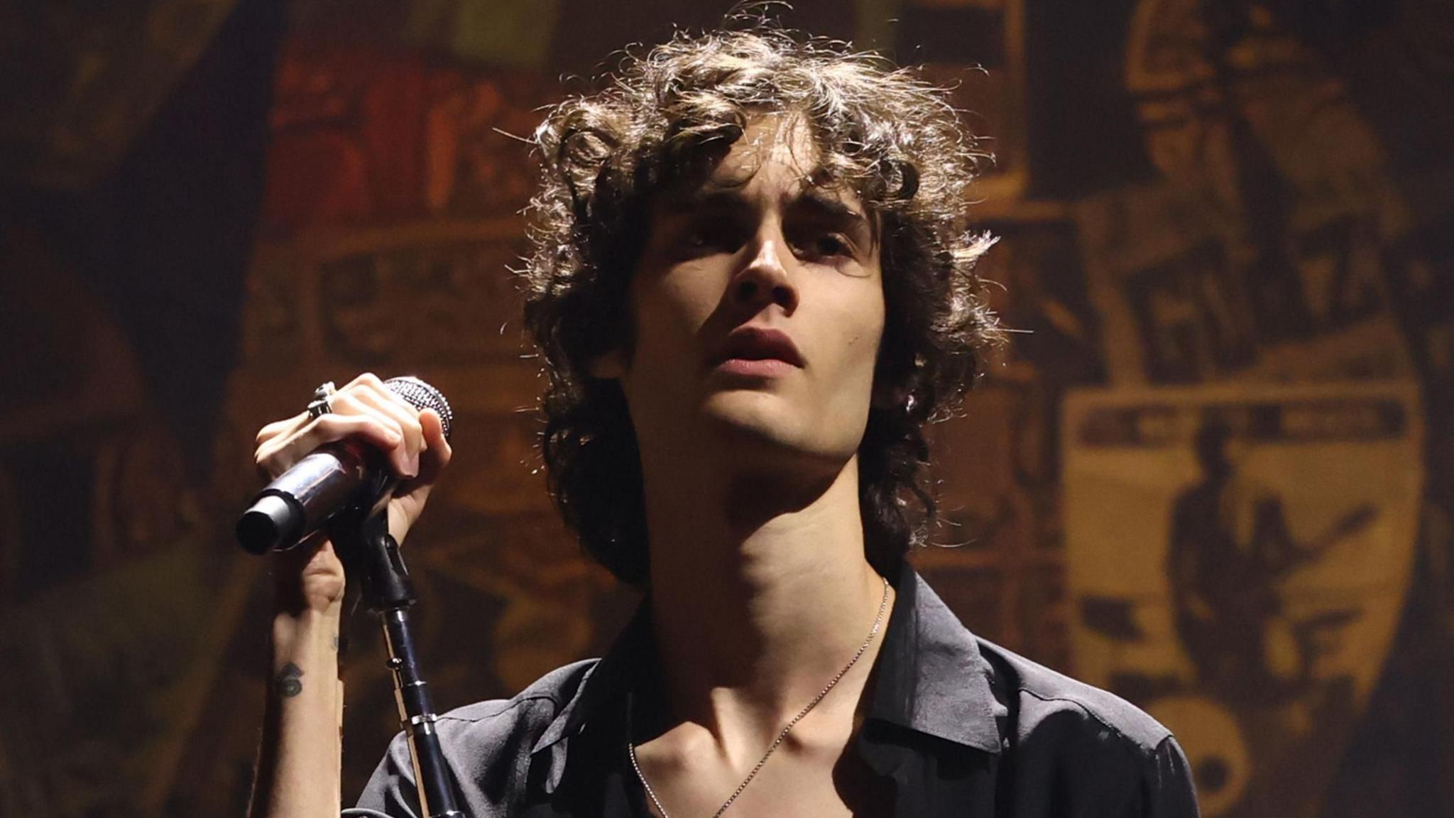 Sombr in a half-unbuttoned black shirt holding a microphone stand and looking moody on stage at the 2025 MTV Video Music Awards