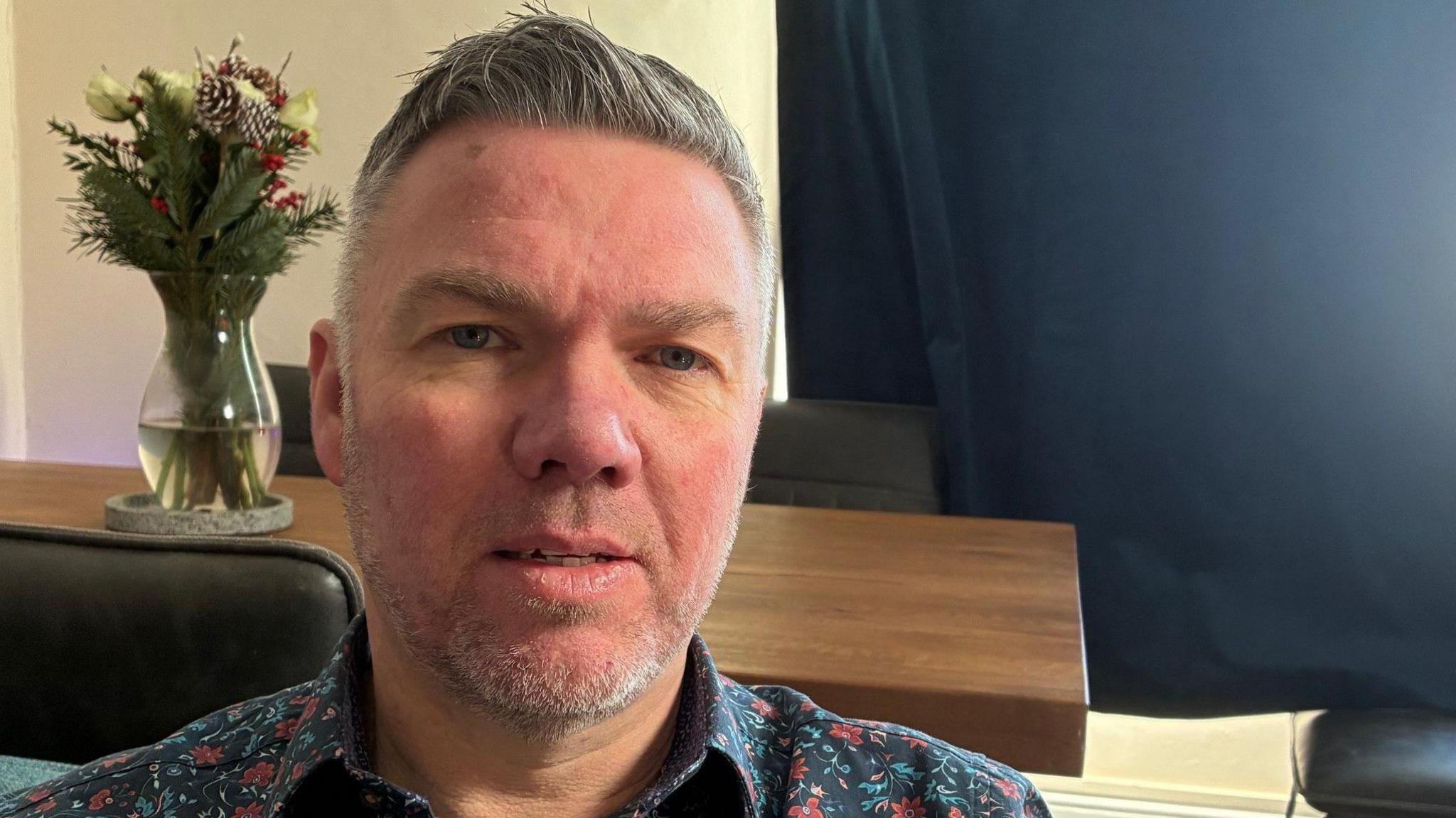 Dave Murphy in a floral shirt sits in front of a table with a vase of flowers on it.