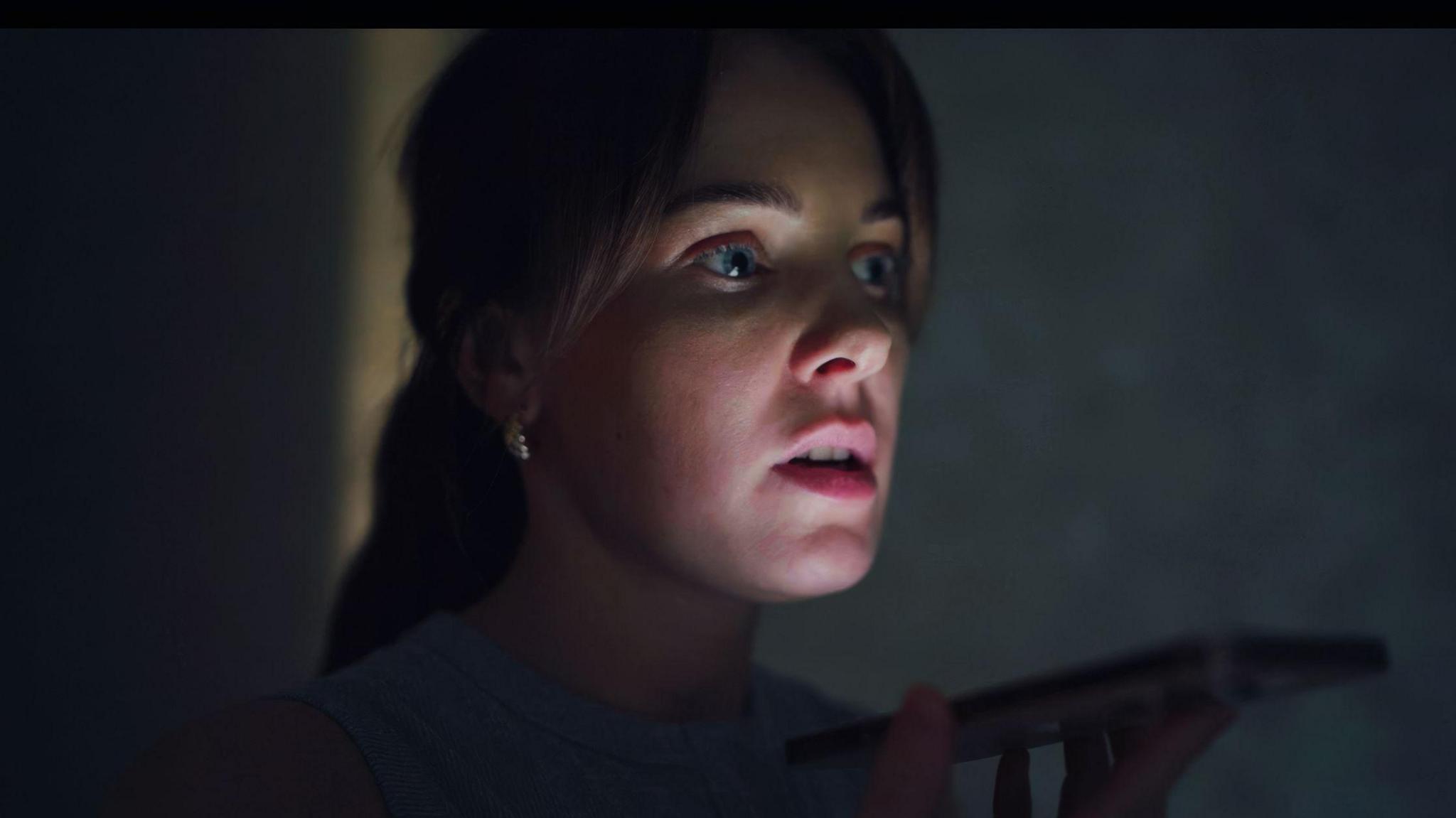 A woman holding a smartphone close to their mouth, wearing a sleeveless grey top, in a dimly lit indoor setting