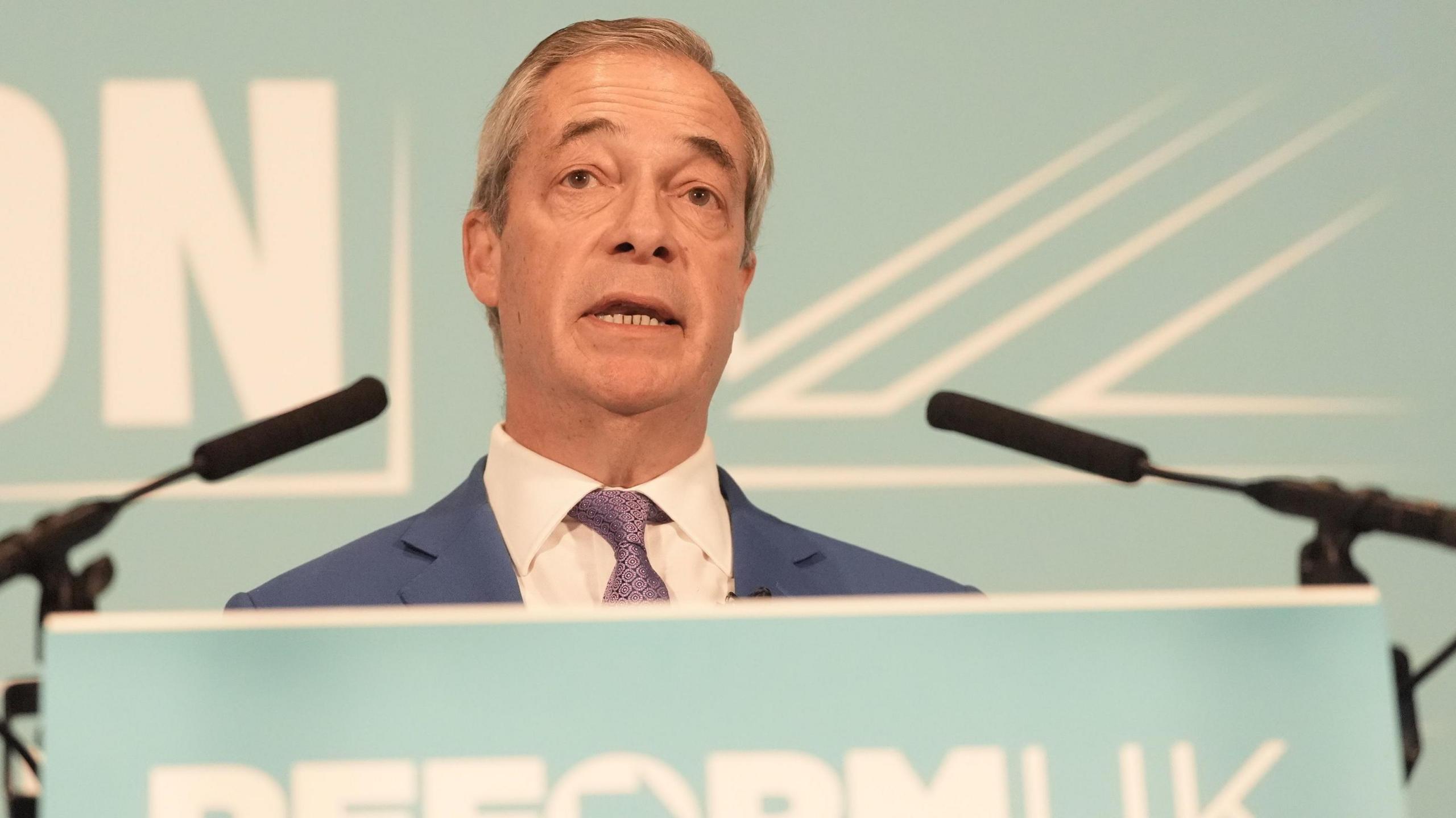 Nigel Farage standing at a branded Reform party lectern