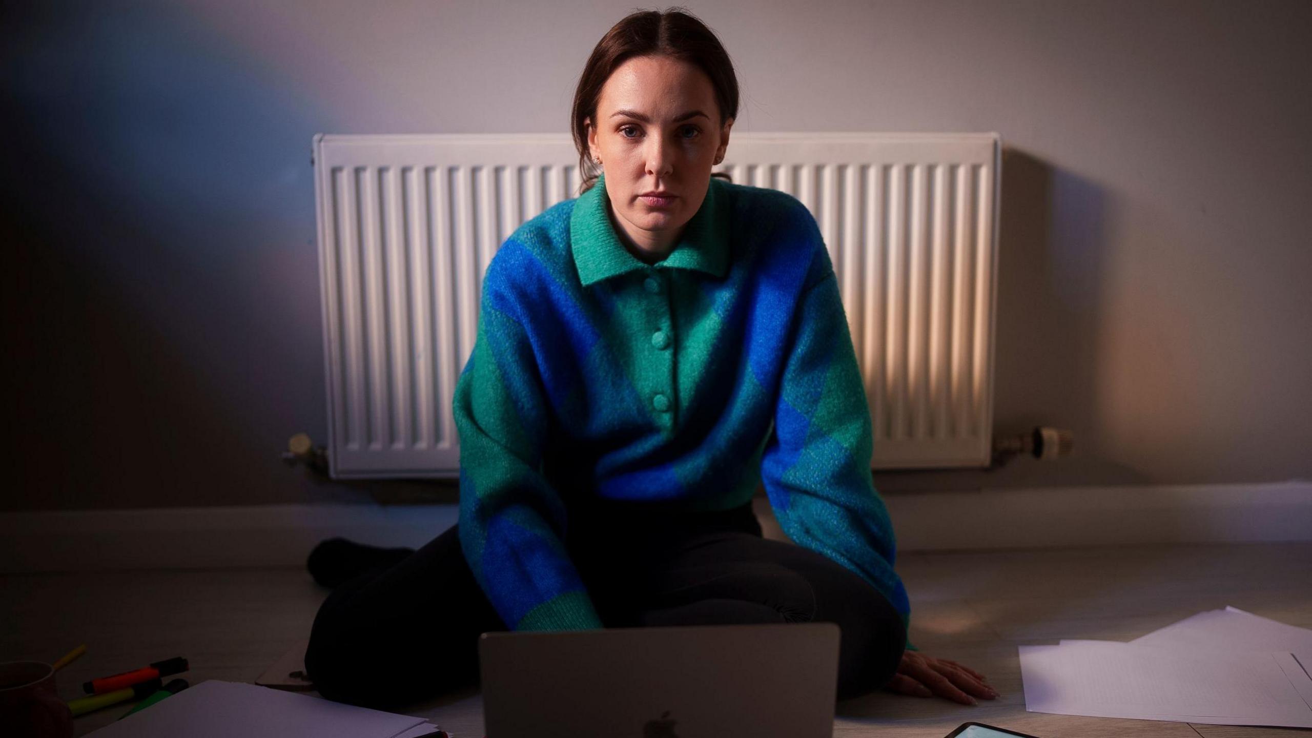 A woman sitting on the floor in front of a white radiator, wearing a blue and green patterned sweater, with a laptop and scattered papers around