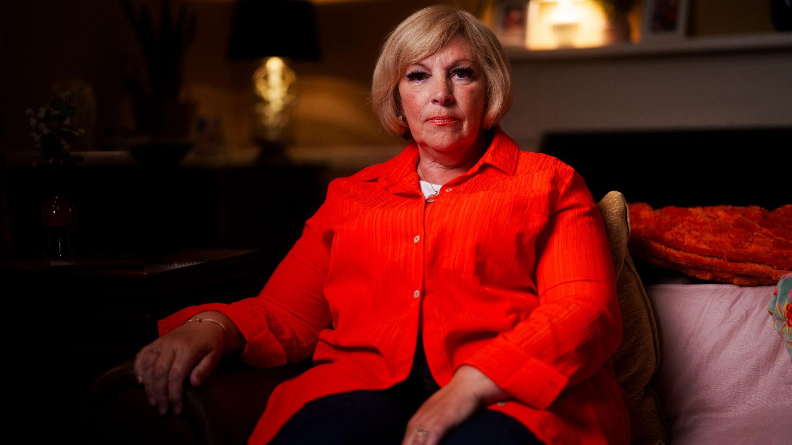 A woman wearing a bright red, button-up shirt, seated indoors on a sofa with a cushion and throw blanket, in a warmly lit living room setting