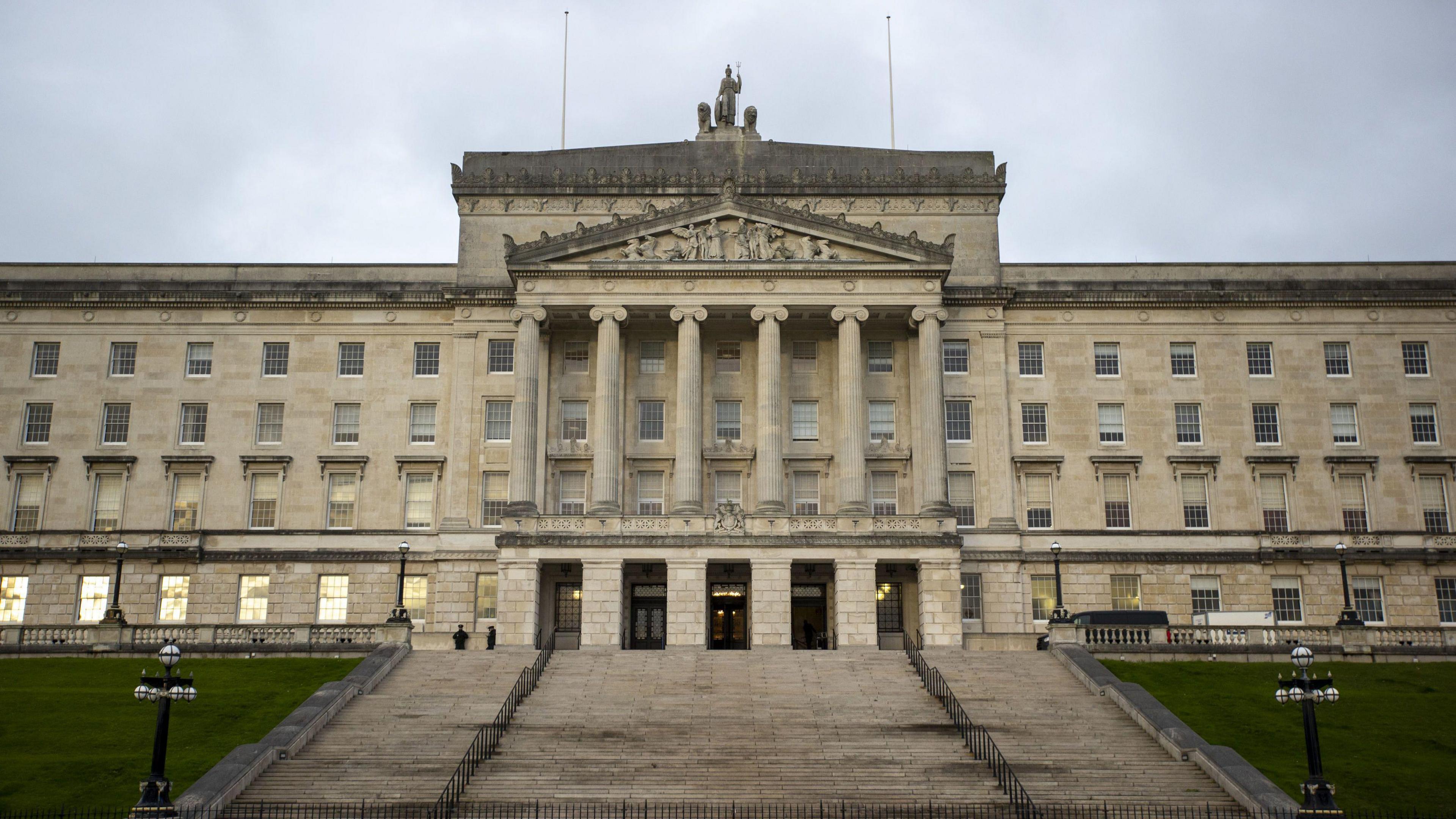 Stormont building from the front. The building is an off white colour. It has six columns on its front and steps leading to the doors. There is grass on either side of these steps.
