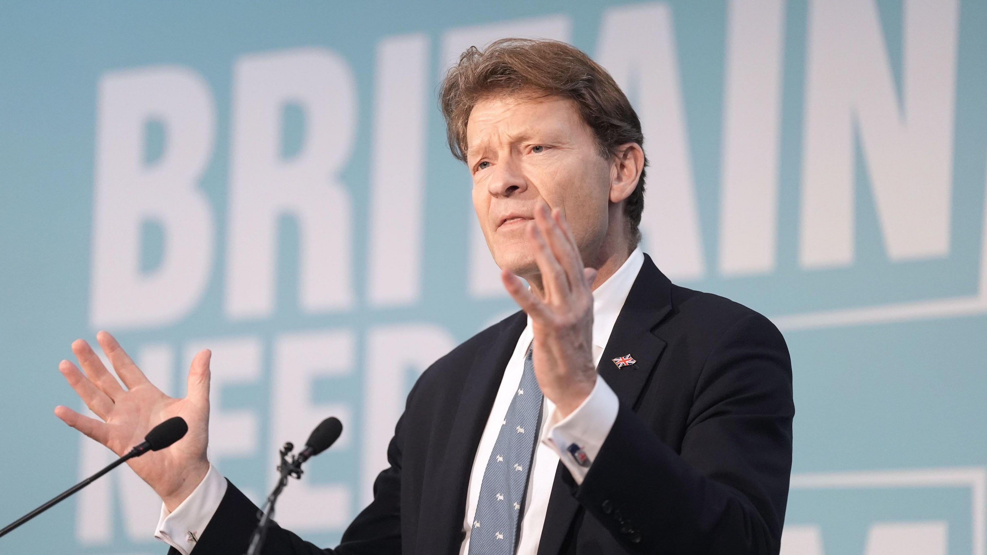 Richard Tice, wearing a black suit jacket and a union flag lapel badge on his left lapel, is talking to an audience with his hands widened.