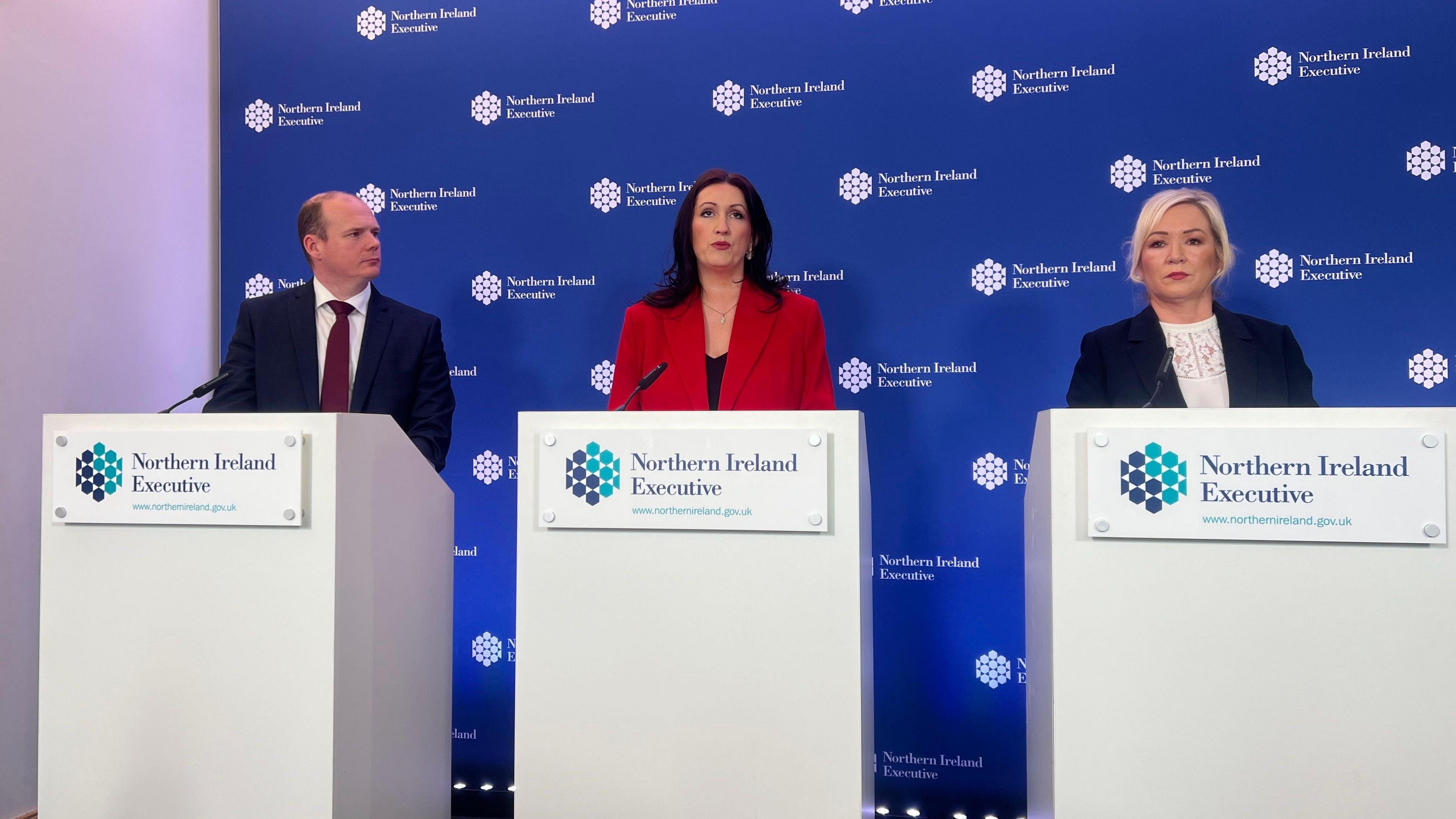 Three people standing at press podiums. A man in a suit, Gordon Lyons, is standing to the left. Emma Little Pengelly - who has longer dark brown hair - in a red suit is to his right and Michelle ONeill - who has shoulder-length blonde hair- in a black suit is toher right. The background is blue and has NI executive logos across it.