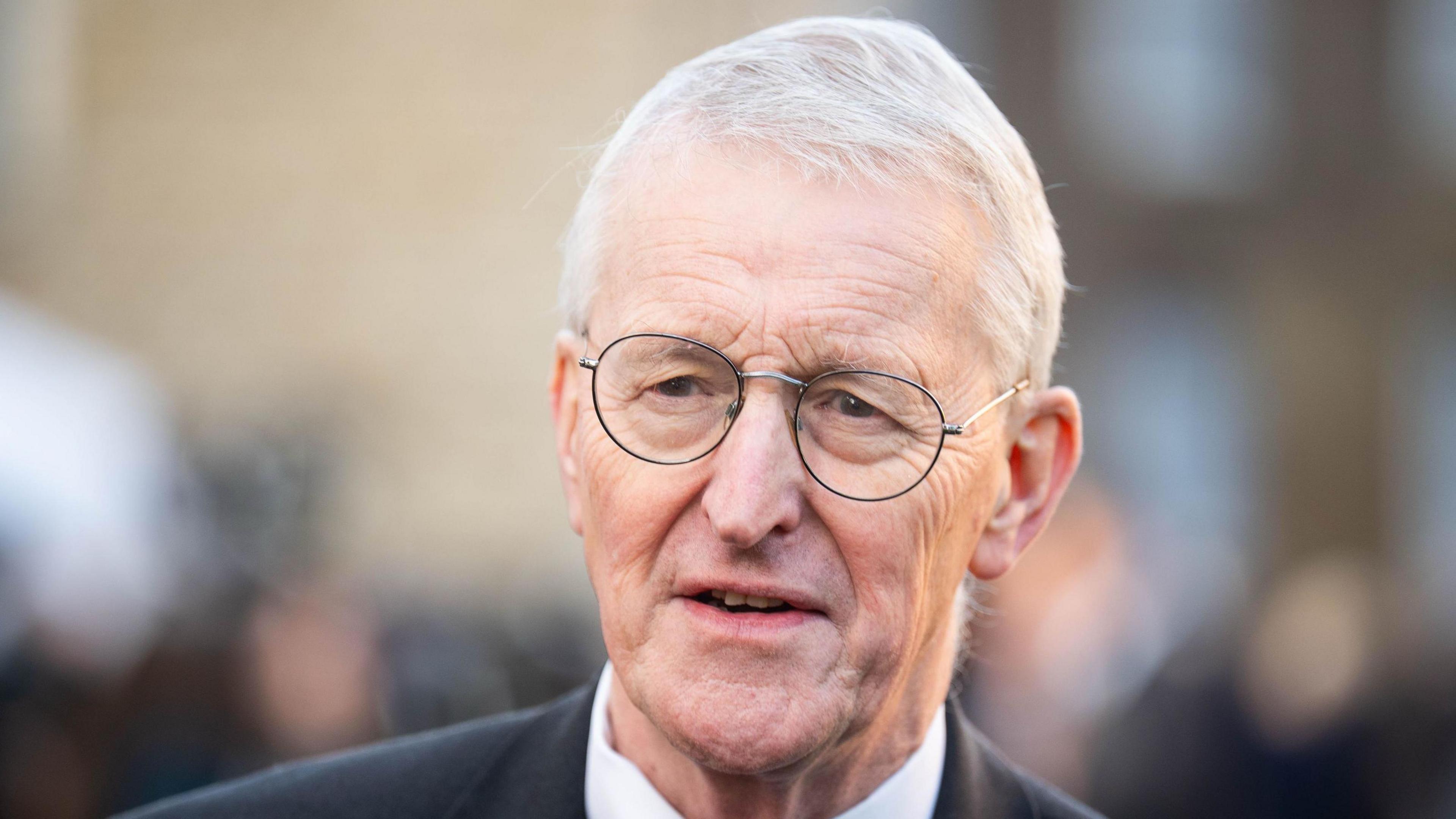 Hilary Benn has short grey hair and thin black frames glasses and is wearing a dark suit.