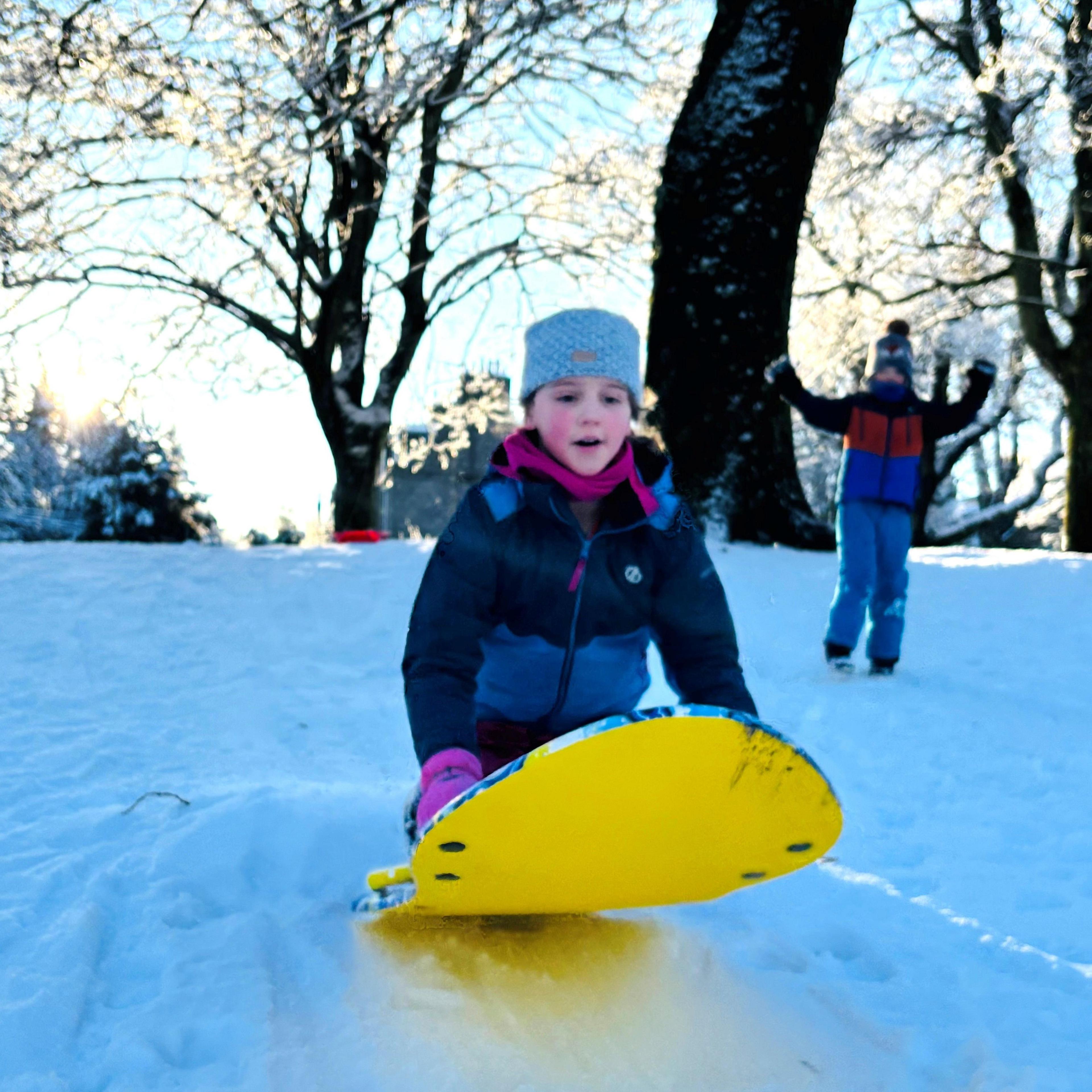 Young girl on a yellow snowboard in the snow with a young boy in the background.