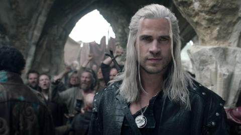 Liam Hemsworth as Geralt with black leather armour on and a sword holstered on his back. He has a silver pendant and long white hair. He stares into the distance. Behind him is a group of angry people in a medieval style area.
