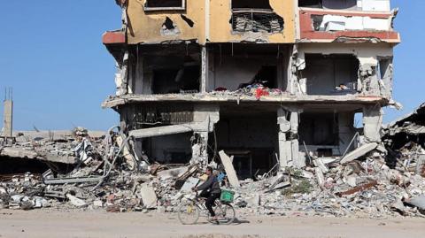 Rubble and a destroyed building as a man on his bike rides past it on a dirt road.
