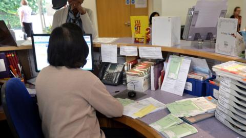 A GP surgery reception. The receptionist has her back to the camera and is talking to a female patient who is standing at the desk. The desk has a phone, PC, prescriptions and filing systems. Beyond is a waiting room where two women are sitting.
