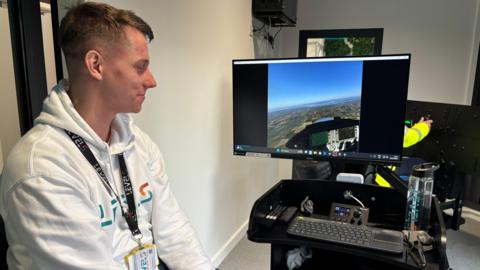 A blond-haired man in a white hoodie is looking at a computer screen.
