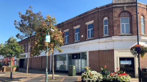 A brick-built, two-storey building in a town centre. There are pedestrians walking down the side of the building and there are raised flower beds in black containers in front of the building.