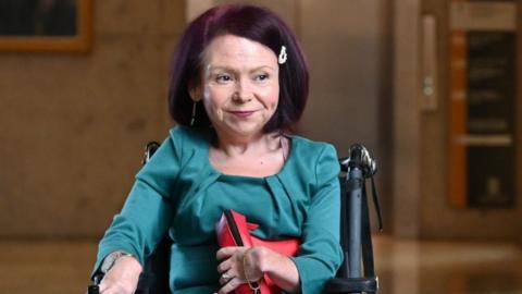 Pam Duncan-Glancy, who has shoulder-length red hair, looks off to the left of the camera. She is wearing a turquoise white top and is in a wheelchair.