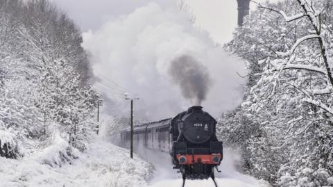 Steam train moving through snow-covered track lined with snow-covered trees