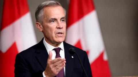 Canada's Prime Minister Mark Carney speaks during a press conference, on the sidelines of the 47th ASEAN Summit in Kuala Lumpur, Malaysia, October 27, 2025. He is wearing a black suit with a white collard shirt and a deep burgundy tie, and is standing in front of two Canadian flags on poles.