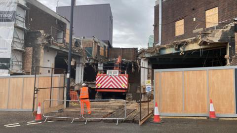 A site entrance that is partially boarded. Behind hoardings machinery is pulling down an old department store. One worker stands in the foreground at the entrance to the site.