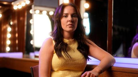 A woman wearing a sleeveless, yellow textured top, seated in front of a mirror with round vanity lights in a dressing room setting.