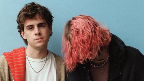 Press photo of Royel Otis standing against a plain blue background. Guitarist Royel Maddell covers his face with long, pink tousled hair