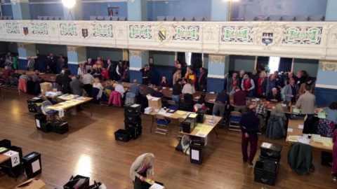 An election count at Oxford Town Hall - the image is taken from a balcony looking down at the hall, where there are counters around trestle tables and black ballot boxes.