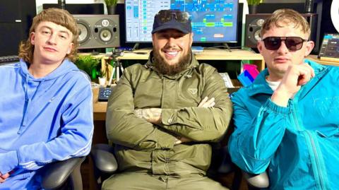 Bad Boy Chiller Crew sit facing the camera in their recording studio. They are all wearing tracksuits. Clive's is light blue, GK is in dark green and Kane's is bright blue. GK is wearing dark sunglasses and has his arms folded. Kane is wearing dark sunglasses. Recording studio equipment is visible behind them with a computer screen showing a mix of their music.