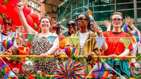 Five people pictured behind a barrier at Manchester Pride. They are waving pride flags smiling and waving. The barrier is decorated with various rainbow flags and disco balls.