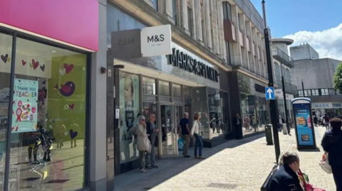 Shoppers are seen leaving a high street store that reads Marks & Spencer above the door. The sun is shining and people are also walking or sitting on seats on the pedestrianised high street outside. The store is next to others shops.