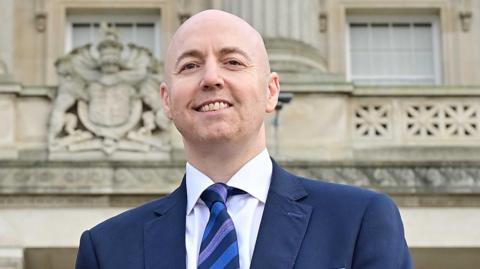 Jon Burrows, is smiling at the camera. He is bald and is wearing a navy jacket over a white shirt and a blue and navy diagonally striped tie. He is standing outside of Parliament Buildings at Stormont in Belfast.