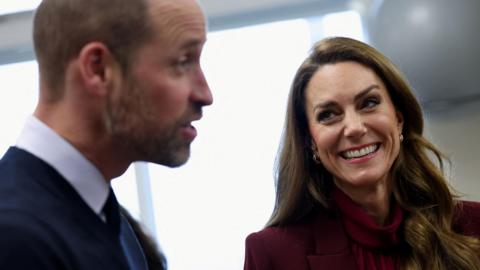 The Prince and Princess of Wales, joint Patrons of NHS Charities Together, during a visit to Charing Cross Hospital, west London. William is pictured to the left of the image and is looking to the right, with his eyebrows raised. Catherine is on the right, smiling at him. Her long brown hair is down and she is wearing a burgundy trouser suit.