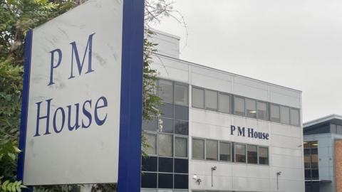 A large office building with the name “PM House” displayed prominently on its exterior. The building has a modern, rectangular design with multiple rows of windows and light grey cladding. In the foreground, a tall sign also reads “PM House” in blue lettering on a white background, framed by a dark blue border. Several cars are parked in front of the building, suggesting a business or office complex.