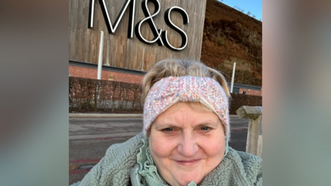 Flick Williams, a woman wearing a green fleece and woolly headband, smiles in front of a large M&S sign.