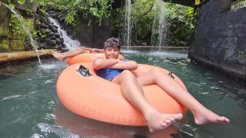 A young boy, with his arms crossed, floating on a inflatable orange ring in a pool with greenery and a waterfall in the background