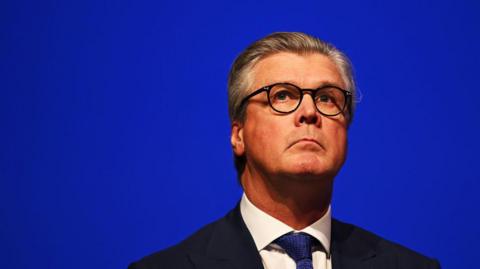 A man with grey hair, black-rimmed round glasses and a dark suit with a white shirt in front of a royal blue background