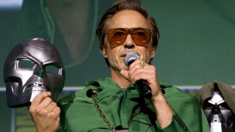 Robert Downey Jr at San Diego's Comic Con event in 2024, when his return to the MCU in a new role was announced on stage. He is wearing the character's recognisable shade of dark green and sunglasses, having just removed Doom's metallic mask, which was hiding his identity from the audience, which he is now addressing using a microphone.