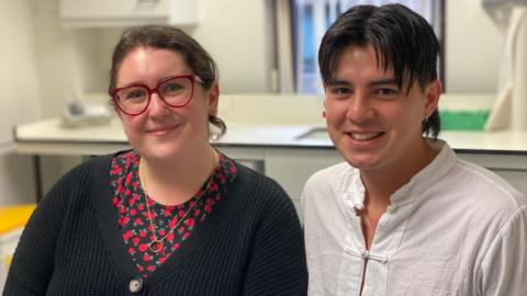 A young woman and a young man sit next to each other, smiling, in a laboratory setting. The young woman wears red-rimmed glasses and a black cardigan over a dark blouse with a red rose pattern. The young man wears a white shirt.