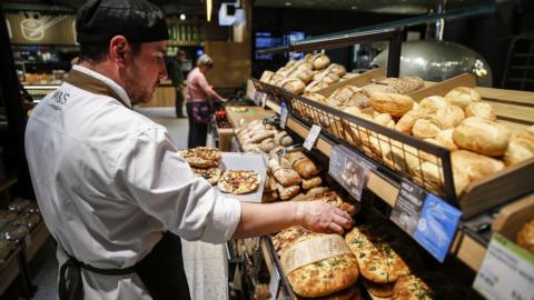 An M&S staff baker restocks the shelves full of pizza and bread rolls