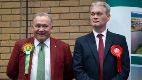 Two men - one smiling with a green Plaid Cymru badge and one not smiling with a red Labour badge - stand on a stage next to each other.