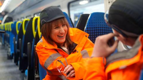 Rachel Reeves sat aboard a train wears an orange hi-viz jacket and a black hard cap.