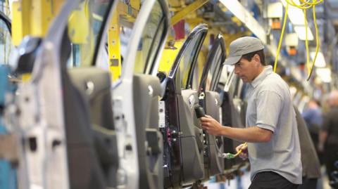 A man in a grey tshirt and cap inspects a row of grey car doors inside a factory.