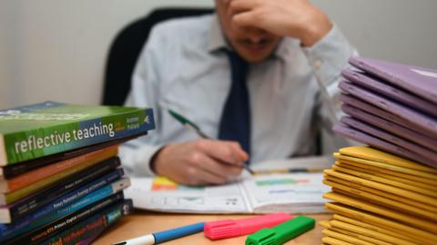 A teacher sits at a desk with their head in their hand, appearing stressed while marking work. Open exercise books and colourful highlighters are on the table. Piles of folders and a stack of teaching guides, including “Reflective Teaching,” surround them.