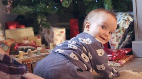 A baby is on his hands and knees on a rug indoors, looking towards the camera, with a decorated Christmas tree and wrapped presents visible in the background.
