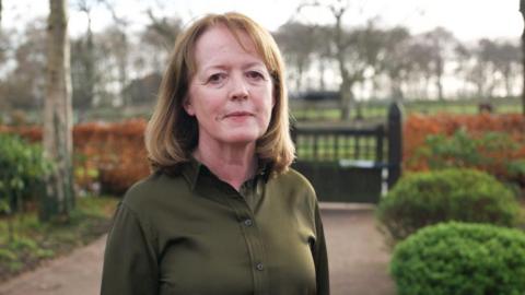 Dr Susan Gilby wears a dark green shirt and is standing on a path in front of a gate with red flowers and tall trees in the background