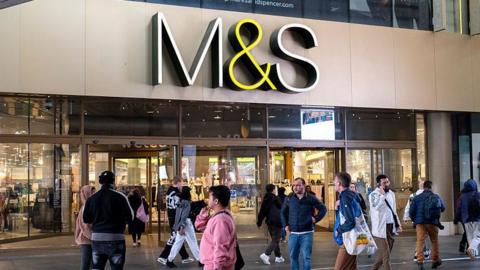 A crowd of people walk outside of a large M&S store with an M&S sign hanging overhead